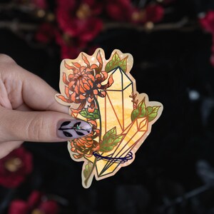 May include: A die-cut sticker featuring a vibrant orange chrysanthemum flower intertwined with two yellow, geometric-shaped crystals. The design includes green leaves and a thin, dark blue wire-like accent wrapped around the base of the crystals.