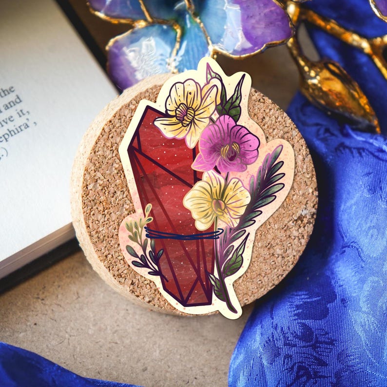 Puede incluir: Una pegatina que presenta un cristal geom&eacute;trico de color rojo intenso adornado con flores amarillas y moradas y follaje verde. El dise&ntilde;o est&aacute; centrado sobre una superficie circular de corcho, mostrando l&iacute;neas precisas y colores vivos.