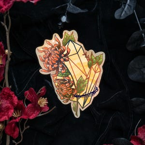 May include: A die-cut sticker featuring a cluster of two orange chrysanthemum flowers intertwined with two yellow geometric crystals and green leaves. The design includes a thin dark blue circular line accenting the base of the crystals.