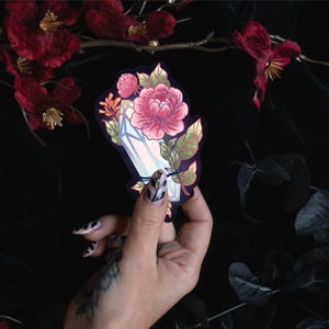 May include: A die-cut sticker featuring a vertical, light-reflecting crystal shard intertwined with a large, detailed pink peony flower and green leaves. The illustration is set against a dark background, showcasing a botanical and mineral design.