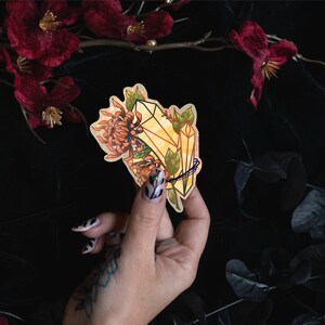 May include: A die-cut sticker featuring a yellow crystal cluster intertwined with orange chrysanthemum flowers and green leaves. The illustration has a warm color palette with defined dark outlines, showcasing a botanical and mineral design.