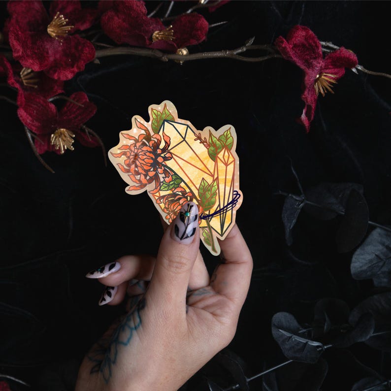 May include: A die-cut sticker featuring a yellow crystal cluster intertwined with orange chrysanthemum flowers and green leaves. The illustration has a warm color palette with defined dark outlines, showcasing a botanical and mineral design.