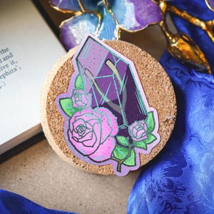 Puede incluir: Una pegatina troquelada que presenta un fragmento de cristal morado superpuesto con dos rosas rosadas y hojas verdes. El diseño tiene una textura sutilmente brillante, con tonos morados oscuros y claros en el cristal y pétalos de color rosa suave.