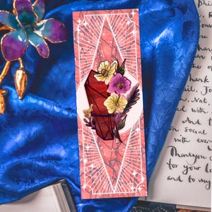 Puede incluir: Un marcapáginas rectangular con fondo color coral y un patrón geométrico de diamantes blancos. En el centro, una ilustración de un cristal rojo oscuro adornada con flores amarillas y moradas, hojas verdes y tallos oscuros finos.