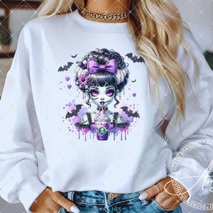 May include: White sweatshirt featuring a spooky cartoon girl with purple and black accents, drinking from a purple cup. The design includes bats, hearts, and floral elements. A fun, Halloween-themed graphic.