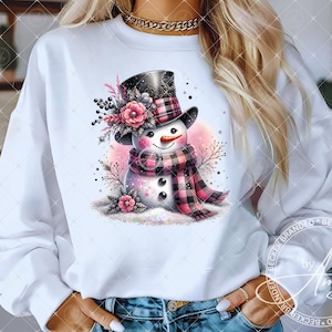 May include: White sweatshirt with a pink and black plaid snowman graphic. The snowman is wearing a black top hat with a pink and white flower design. The snowman is surrounded by pink and white flowers and green leaves.