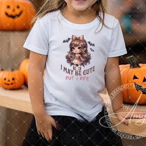 May include: A white t-shirt with a cute cartoon girl wearing a pink dress and holding a candy corn. The text on the shirt reads "I may be cute but I bite".
