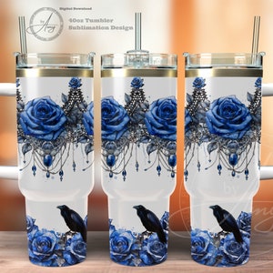 Op de afbeelding: Een witte 40oz tumbler met een gouden deksel en een zwart kraaienontwerp. De tumbler heeft een blauw rozen- en zilveren sieradenontwerp. De tumbler staat op een houten oppervlak.