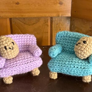 May include: Two crocheted armchairs in purple and blue with a crocheted person sitting in each chair. The person in the purple chair is smiling and the person in the blue chair is looking to the side.