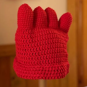 Puede incluir: Un gorro de ganchillo rojo brillante, con forma de mano con los dedos hacia arriba. El gorro está hecho de un hilo grueso y texturizado. Este gorro de novedad es probablemente para disfraces o uso lúdico.