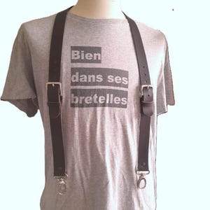 Peut inclure: Un t-shirt gris à manches courtes portant le texte Bien dans ses bretelles en lettres gris foncé, porté avec des bretelles en cuir marron foncé dotées de boucles en métal argenté et de fermoirs mousquetons pivotants à l'avant.