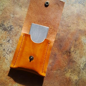 Può includere: Un fodero in pelle fatto a mano, aperto, in una calda tonalità marrone dorato, con un oggetto metallico argentato parzialmente visibile all'interno. Il fodero è assicurato con una chiusura a scatto e presenta cuciture dettagliate lungo i bordi. La pelle ha un aspetto naturale e strutturato.
