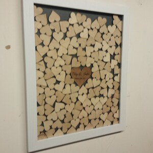 Op de afbeelding: Een wit frame met een grijze achtergrond gevuld met houten hartvormen. De harten zijn in een patroon gerangschikt en er is een groter hart in het midden met de tekst "Clay & Justin" en de datum "1 oktober 2022".