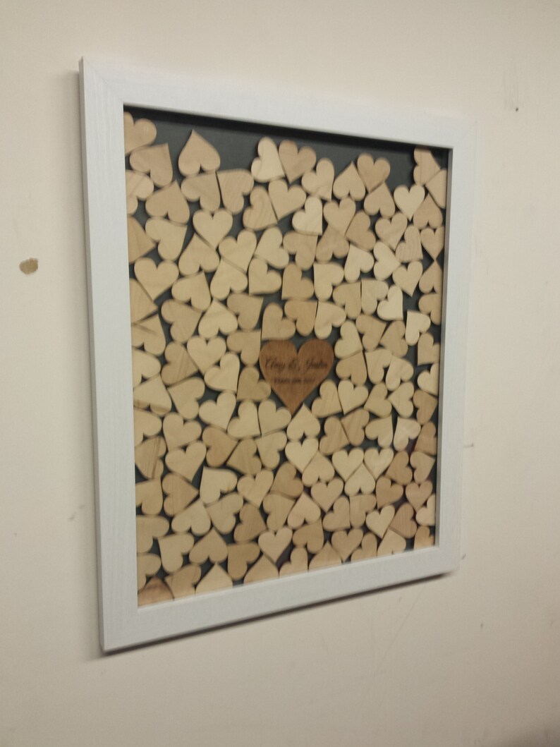 Op de afbeelding: Een wit frame met een grijze achtergrond gevuld met houten hartvormen. De harten zijn in een patroon gerangschikt en er is een groter hart in het midden met de tekst "Clay & Justin" en de datum "1 oktober 2022".