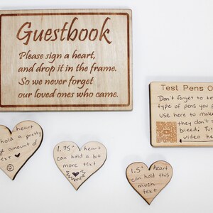 Op de afbeelding: Een houten bord met de tekst "Guestbook" en instructies om een hart te tekenen en in het frame te stoppen. Drie houten harten in verschillende maten zijn te zien met handgeschreven tekst erop. Een kleiner houten bord met de tekst "Test Pens On Me" en instructies om de pennen te testen voordat je ze gebruikt, is ook te zien.