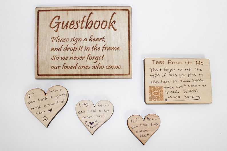 Op de afbeelding: Een houten bord met de tekst "Guestbook" en instructies om een hart te tekenen en in het frame te stoppen. Drie houten harten in verschillende maten zijn te zien met handgeschreven tekst erop. Een kleiner houten bord met de tekst "Test Pens On Me" en instructies om de pennen te testen voordat je ze gebruikt, is ook te zien.