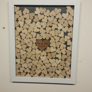 Op de afbeelding: Een wit frame met een grijze achtergrond gevuld met houten harten. Een groter houten hart met de tekst "Philip & Sarah" bevindt zich in het midden van het frame.