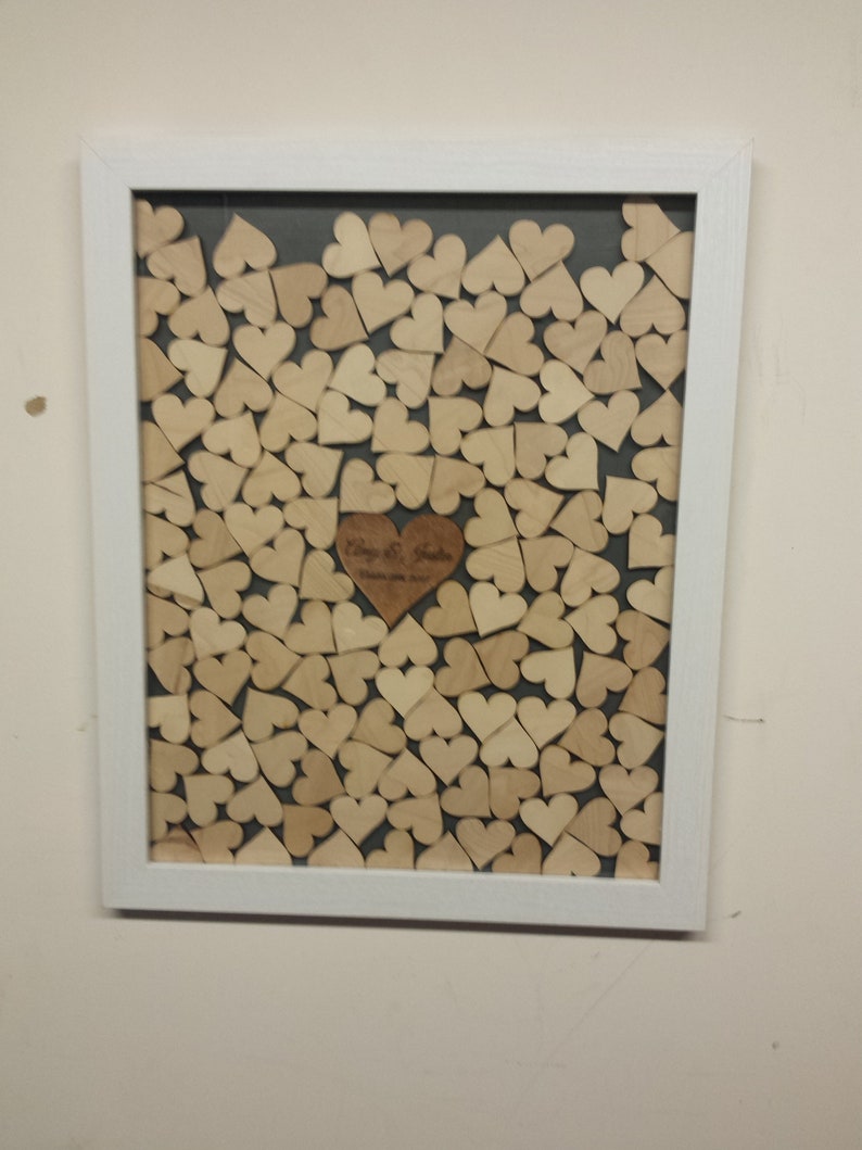 Op de afbeelding: Een wit frame met een grijze achtergrond gevuld met houten harten. Een groter houten hart met de tekst "Philip & Sarah" bevindt zich in het midden van het frame.