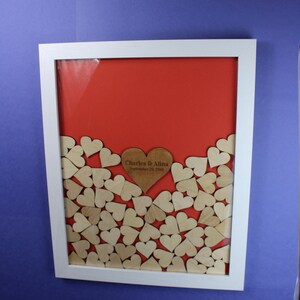Op de afbeelding: Een wit frame met een rode achtergrond en een houten hart met de tekst "Charles & Alina September 29, 2018" omringd door kleinere houten harten.
