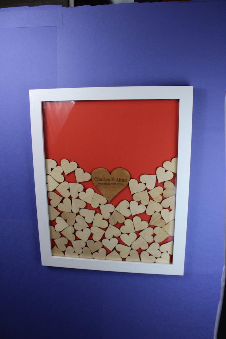 Op de afbeelding: Een wit frame met een rode achtergrond en een houten hart met de tekst "Charles & Alina September 29, 2018" omringd door kleinere houten harten.