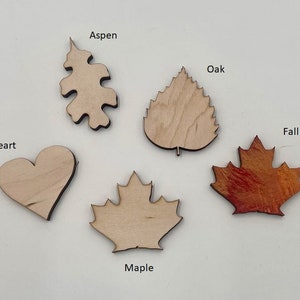 May include: A collection of wooden leaf and heart cutouts. The shapes include an Aspen leaf, an Oak leaf, a Maple leaf, a heart, and a Fall leaf with red and orange tones. Text labels identify each shape.