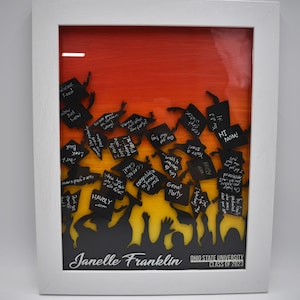 May include: A white frame with a red, orange, and yellow background. Silhouettes of people are holding up black graduation caps with handwritten messages. The text "Janelle Franklin OHIO STATE UNIVERSITY CLASS OF 2023" is at the bottom of the frame.