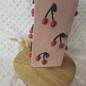 Op de afbeelding: Een rechthoekige, lichtroze keramische vaas met een getextureerde, matte afwerking. Het oppervlak is versierd met meerdere verhoogde, driedimensionale kersmotieven, elk bestaande uit twee kleine rode bolletjes aan zwarte steeltjes in een patroon.