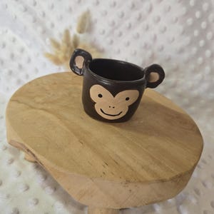 Peut inclure: Une petite tasse en céramique marron foncé en forme de singe. La tasse présente un visage de singe beige clair peint sur le devant, avec deux oreilles saillantes sur les côtés. L'intérieur de la tasse est marron foncé, assorti à la finition extérieure.
