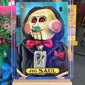 May include: A trading card featuring a character with a pale face, red eyes, and spiral cheeks. The character wears a black suit, red bow tie, and holds a cassette player that says "PLAY ME". The card is labeled "JIG SAUL".