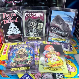May include: A collection of Garbage Pail Kids and other trading cards, including "Feces of Death," "Crap Show," and "Tanked Frank." The cards are in protective sleeves, with colorful illustrations and text. Boxes of Batman cards are visible.