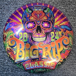 May include: A colorful disc golf disc with a skull design. The disc features a vibrant skull with floral accents, mushrooms, and the text "2024 BIG RIP CLASSIC". The disc is primarily purple, orange, and green.