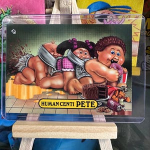 May include: A trading card featuring three cartoon babies with a green background. The babies are wearing diapers and are eating junk food. The card has the text "HUMAN CENTI PETE" on it.