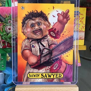 May include: A yellow and red trading card featuring a cartoon illustration of a doll holding a chainsaw. The doll is wearing a white shirt with a red stain and a blue bandana. The text "SAWIN' SAWYER" is printed on the bottom of the card.