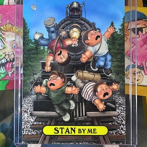 May include: A trading card featuring a cartoon illustration of four children in distress, seemingly about to be hit by a train. The card has the text "STAN BY ME" in a yellow banner. The background shows a train and a forest.