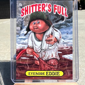 May include: A collectible trading card featuring a cartoon character with a red face, green hat, and white robe. The card has the text "SHITTER'S FULL" in red and "EYESORE EDDIE" in yellow.