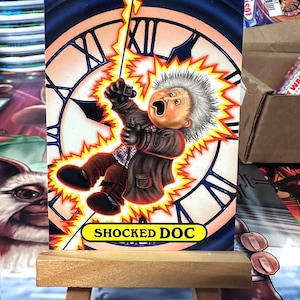 May include: A trading card featuring a cartoon character with white hair and a shocked expression. The character is holding a device and is surrounded by yellow and orange lightning bolts. The card has a blue background with a clock face and the text "SHOCKED DOC".
