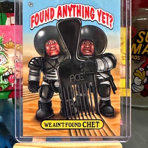 May include: A trading card featuring a cartoon illustration of two black figures wearing black helmets and holding a black comb. The text on the card reads "FOUND ANYTHING YET?" and "WE AIN'T FOUND CHET".