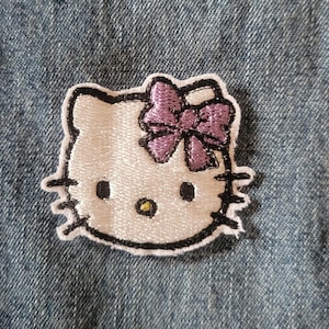 May include: Embroidered patch featuring the iconic Hello Kitty character. The patch has a white face with black eyes, whiskers, and a purple bow. The patch is outlined in black and is set against a blue denim background.