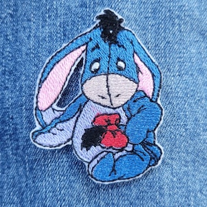 May include: Embroidered patch featuring Eeyore from Winnie the Pooh. The patch depicts Eeyore in shades of blue, with pink inner ears, a gray muzzle, and a red bow. The patch is on a denim background.