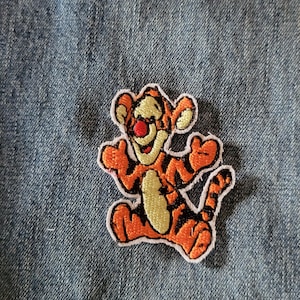 May include: Embroidered patch featuring Tigger from Winnie the Pooh. The patch has a white border and depicts Tigger in orange, black, and yellow. The patch is on a blue denim background.