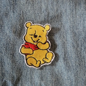 May include: A yellow Winnie the Pooh patch with a red scarf and black outline. The patch is embroidered with a smiling Pooh Bear design. The patch is on a blue denim background.