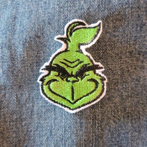 May include: Embroidered patch featuring the Grinch's face in green, black, and white. The Grinch's face is centered, with a green face, black eyebrows, and a white border. The patch is on a blue denim background.