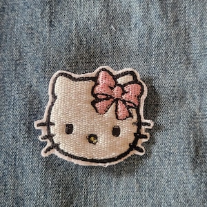 May include: Embroidered patch featuring the iconic Hello Kitty character. The patch has a white face with black eyes, whiskers, and a pink bow. The patch is outlined in black and is set against a blue denim background.