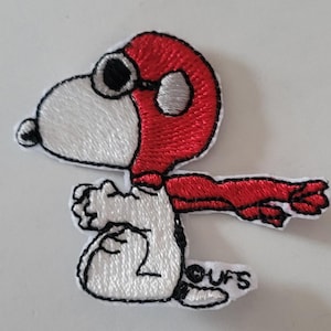 May include: Embroidered patch featuring Snoopy in a red aviator helmet and scarf, with black goggles. The patch is white with black outlining and the text "OUFS" at the bottom. The design depicts Snoopy in a flying pose.