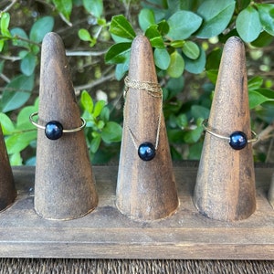 May include: Three gold rings with black pearls on display on wooden ring holders. The rings are simple and elegant.