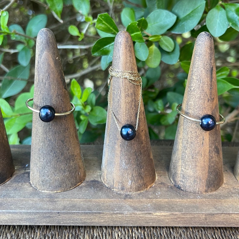 May include: Three gold rings with black pearls on display on wooden ring holders. The rings are simple and elegant.