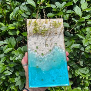 Puede incluir: Una escena de playa en miniatura con una valla de madera, musgo verde y arena blanca. El océano está representado por resina azul con cuentas de vidrio verde.