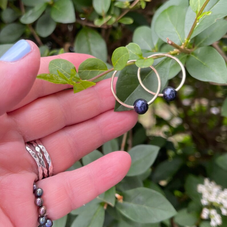May include: A pair of silver hoop earrings with black pearl accents. The earrings are held in a hand with a few other rings on the fingers.