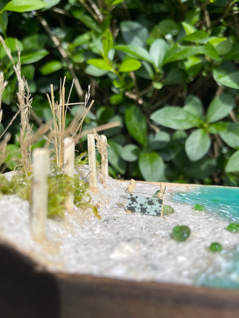 May include: A miniature beach scene with a small wooden fence, green moss, and a tiny beach towel with a black and white pattern. The scene is set in a wooden frame with a blue resin ocean.