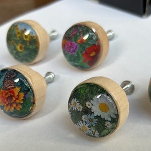 May include: A collection of wooden cabinet knobs with floral designs. Each knob features a different flower, including sunflowers, roses, daisies, and irises, set against a clear, glossy resin. The knobs have silver-toned metal stems.